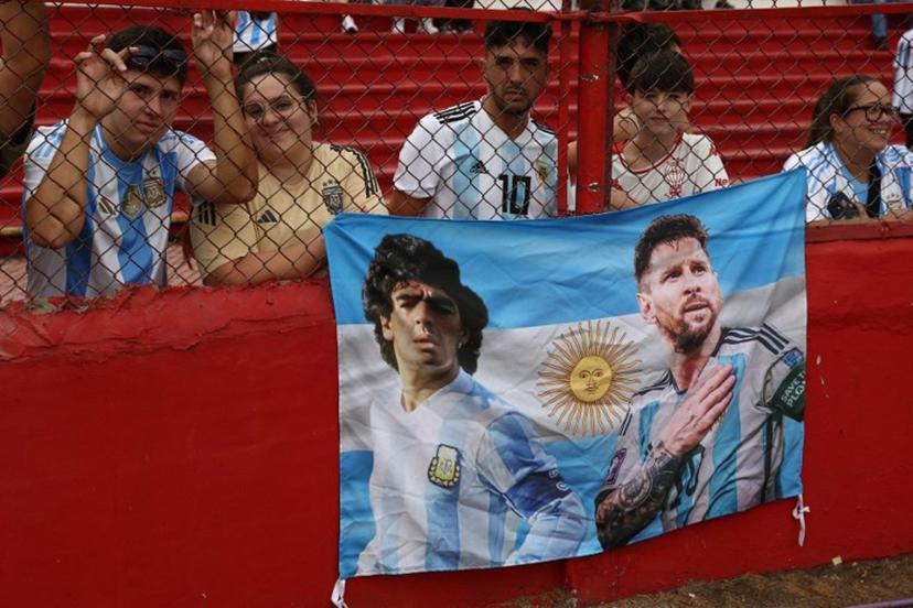 Argentina's national football fans with a flag with the picture of late Argentine football legend Diego Maradona and Forward Lionel Messi attend a training session with the U20 national team at the Tomas Adolfo Duco Stadium in Buenos Aires, on March 22, 2025, in support of the city of Bahia Blanca, hit by a storm that left 16 people dead. Argentina will face Brazil on March 25 in the South American qualifiers for the FIFA 2026 World Cup. ALEJANDRO PAGNI / AFP